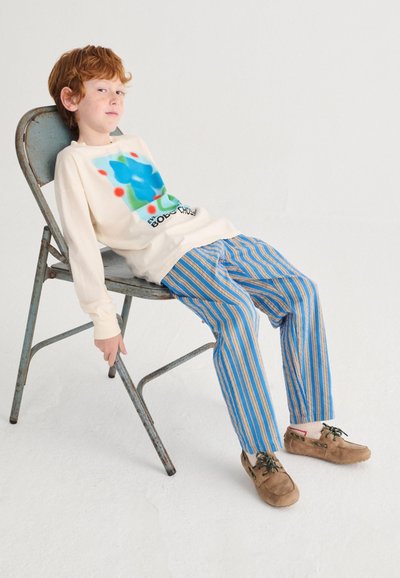 Red-haired child in cream graphic sweatshirt and blue striped pants leans back on a metal chair against a plain white background.