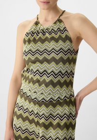 Knit halter top featuring a zigzag pattern in olive green, black, white, and light pink. Soft texture with a gathered neckline and open sides.
