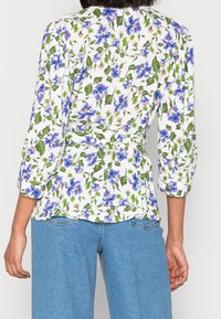 Blusa floreale con sfondo bianco, caratterizzata da fiori blu e foglie verdi. Maniche a tre quarti e design con vita cinte.