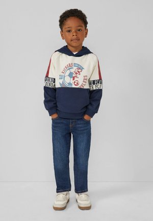 Blauwe en crèmekleurige hoodie met dinosaurussen en voetbalgraphics, rode accentmouwen, gecombineerd met donkere spijkerbroek en witte sneakers.