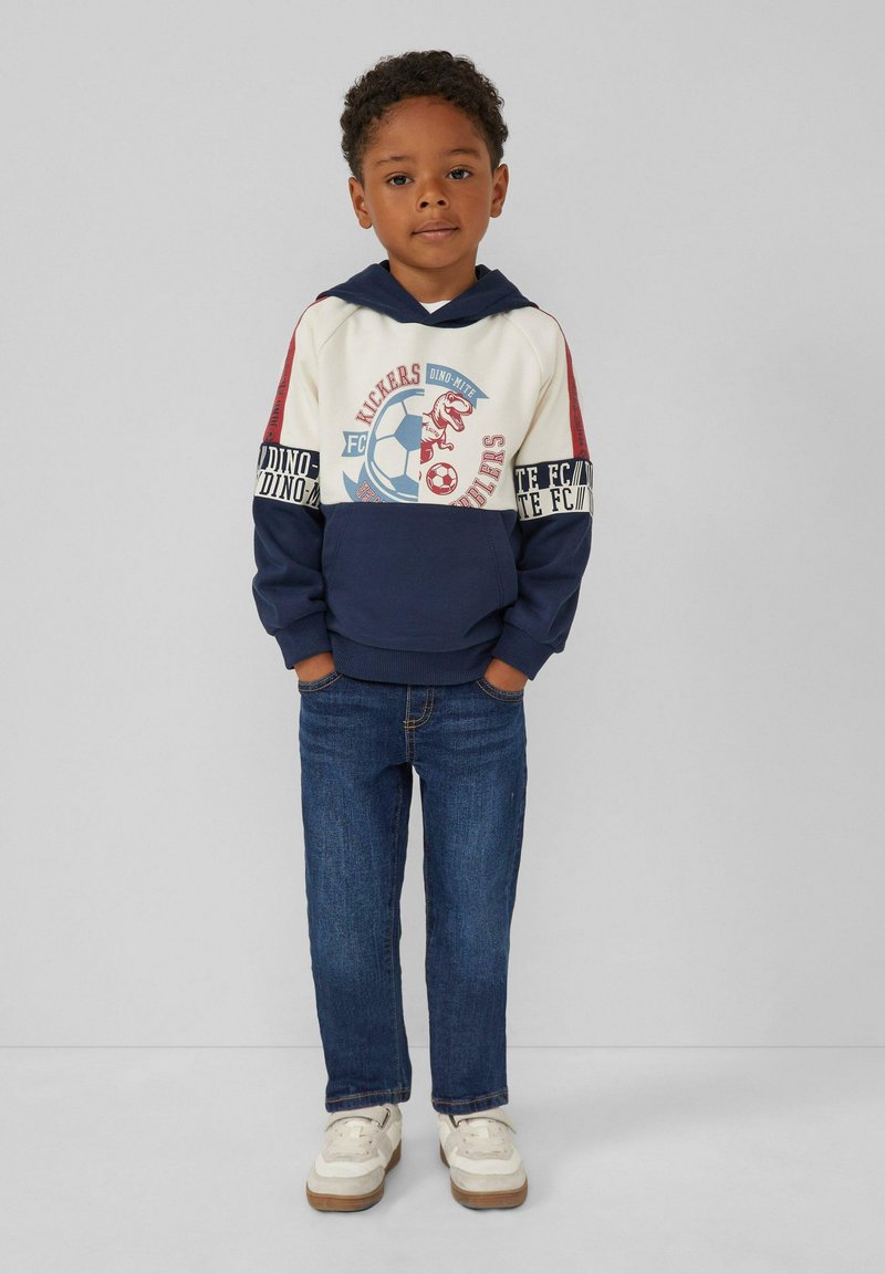 Hoodie bleu et crème avec des graphiques de dinosaure et de football, manches rouges, associé à un jean en denim foncé et des baskets blanches.