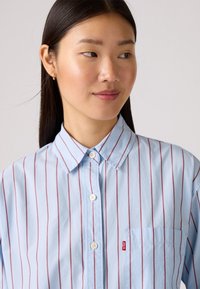 Chemise bleu clair à rayures verticales rouges, avec un col et une poche poitrine. Présente une petite étiquette de logo sur la poche.