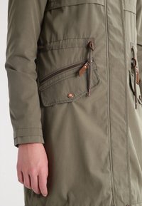 Olivově zelená parka bunda se zapínáním na zip, pasem na stažení a hnědými koženými akcenty. Hladká textura a středně dlouhý střih.