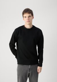 Crna dukserica s okruglim izrezom, dugim rukavima i rebrastim manžetama. Ima suptilan logo na prsima. Uparena s sivim trapericama.