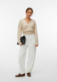 Femme portant un pull beige à col en V, un pantalon blanc taille haute à jambes larges, des chaussures marron, tenant une pochette noire, debout devant un fond blanc.