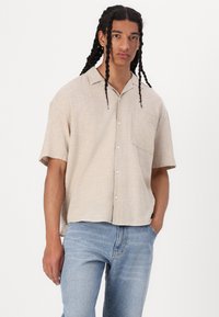 Chemise à manches courtes, de couleur beige clair, avec une poche avant, un ourlet arrondi et une texture subtile. Associée à un jean en denim bleu clair.