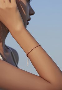 Pulsera de cadena retorcida en oro rosa con un delicado detalle de cadena colgante, llevada en un brazo bronceado contra un fondo de cielo azul claro.