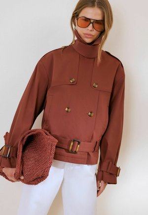 Kvinde iført oversized brun jakke med store knapper og bælte, holder en matchende tekstureret taske og bærer store brune solbriller.