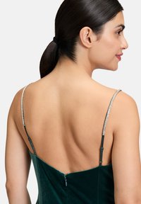 Zielona aksamitna sukienka o niskim dekolcie z tyłu. Cienkie ramiączka ozdobione są kryształowymi akcentami. Tkanina ma miękką, bogatą teksturę i gładkie wykończenie.