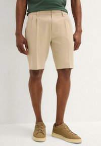 Pantaloni corti beige su misura con pieghe, realizzati in un materiale leggero, abbinati a sneakers color nocciola. I pantaloni corti presentano una chiusura con bottone e cerniera.