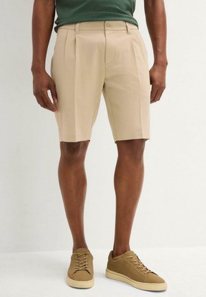 Shorts beige ajustés avec des plis, fabriqués dans un matériau léger, associés à des baskets beige. Les shorts présentent une fermeture à bouton et à glissière.