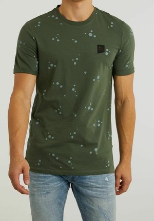 Mann trägt olivgrünes kurzärmeliges T-Shirt mit hellblauem Sprenkelmuster und hellblauen, abgenutzten Jeans, steht vor schlichtem Hintergrund.