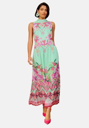 Kvinde iført en ærmeløs mintgrøn maxikjole med pink blomsterprint og klare pink hæle, stående mod en ensfarvet baggrund.
