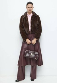 Geacă scurtă din blană artificială maro, cămașă roz deschis, fustă din piele burgundă și pantaloni largi, cizme negre cu toc, poartă o geantă mică maro.