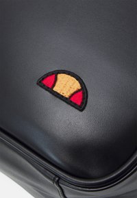 Czarna skórzana torba z małym haftowanym logo o żółtym środku i czerwonych półokręgach, podkreślająca fakturę i szczegóły designu.