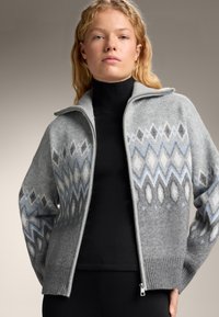 Šedý svetr na zip se vzorem modrých a bílých diamantových tvarů, s žebrovaným lemem a měkkým, texturovaným povrchem.