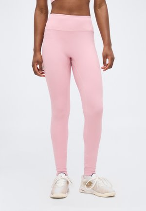 Γυναίκα που φοράει ροζ κολάν με ψηλή μέση και λευκά αθλητικά παπούτσια, στέκεται με τα χέρια χαλαρά δίπλα στο σώμα σε απλό φόντο.