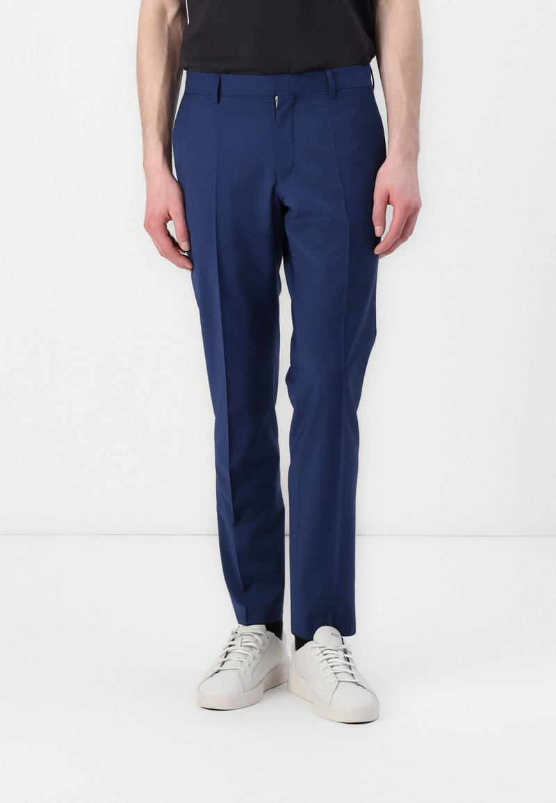 Blaue maßgeschneiderte Hose mit glatter Textur, ausgestattet mit einem Reißverschluss, Gürtelschlaufen und schmaler Passform, kombiniert mit weißen Sneakern.