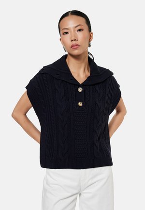 Donna che indossa un maglione nero senza maniche lavorato a trecce con un ampio colletto e due bottoni dorati, abbinato a pantaloni bianchi, mani sui fianchi.