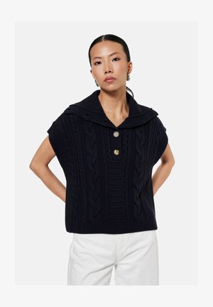 Frau trägt einen ärmellosen schwarzen Kabelstrickpullover mit weitem Kragen und zwei goldenen Knöpfen, kombiniert mit weißen Hosen, Hände in die Hüften gestützt.