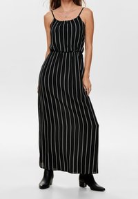 Robe maxi noire à rayures verticales blanches, bretelles spaghetti et taille élastique, associée à des bottines à talons noires.