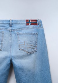 Jeans de mezclilla azul claro con un parche burdeos que muestra la bandera noruega. Cuenta con dos bolsillos traseros con costuras decorativas.
