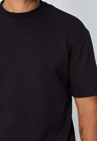 T-shirt noir à manches courtes en mélange de coton, agrémenté de clous métalliques dispersés pour la texture. Col ras de cou classique et coupe décontractée.