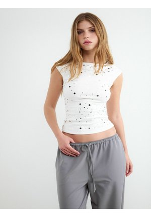 Młoda kobieta z długimi blond włosami, ubrana w biały top bez rękawów z okrągłymi wycięciami oraz szare spodnie ze sznurkiem, stoi na jednolitym tle.