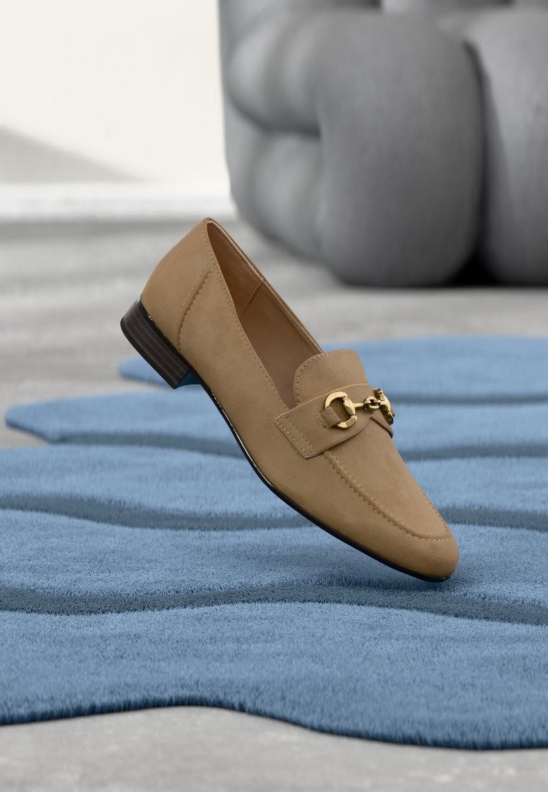 Mocassin en daim marron clair avec un détail de chaîne en or, présenté sur un tapis bleu texturé dans un cadre intérieur moderne.