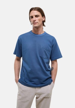Homme aux cheveux bruns aux épaules, portant une chemise bleue à manches courtes avec une poche poitrine et un pantalon beige clair, mains dans les poches.