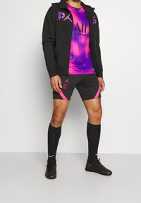 Conjunto deportivo que incluye una sudadera con cremallera negra, una camiseta gráfica vibrante en púrpura y rosa, pantalones cortos negros con acentos rosas y calcetines negros con zapatos rojos.
