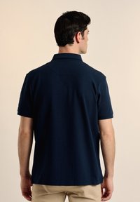 Camiseta polo azul marino de manga corta y cuello clásico, hecha de una tela texturizada. Cuenta con un canesú trasero y un logo bordado en la manga.