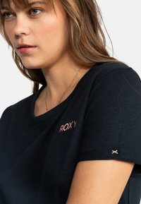 Sort kortærmet T-shirt med V-hals, der har et pink og hvidt "ROXY" logo på brystet samt en lille broderet detalje på ærmet.