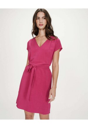 Femme aux longs cheveux bruns portant une robe rose sans manches avec un décolleté en V et une ceinture nouée à la taille, debout devant un fond clair uni.