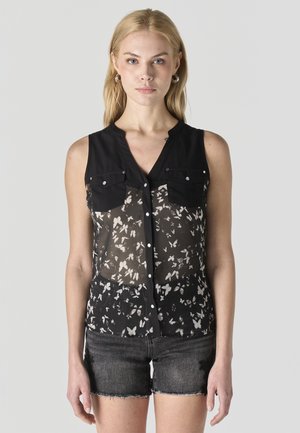 Mujer rubia con blusa negra sin mangas y transparente con estampado de mariposas y pantalones cortos de mezclilla negros con botones, sobre un fondo liso.