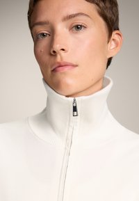 Weißer Reißverschluss-Pullover mit geripptem Textur und hohem Kragen. Verfügt über einen silbernen Reißverschluss mit einem schwarzen Ziehband. Minimalistisches Design.