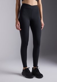 Leggings noirs en tissu extensible, dotés d'une taille haute avec un design en forme de V. Texture lisse, silhouette ajustée.