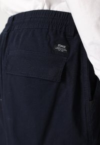 Marina blå shorts med elastisk midja, utrustade med en bakficka och en svart tyglabel med texten "STANCE". Strukturerad tygkvalitet.