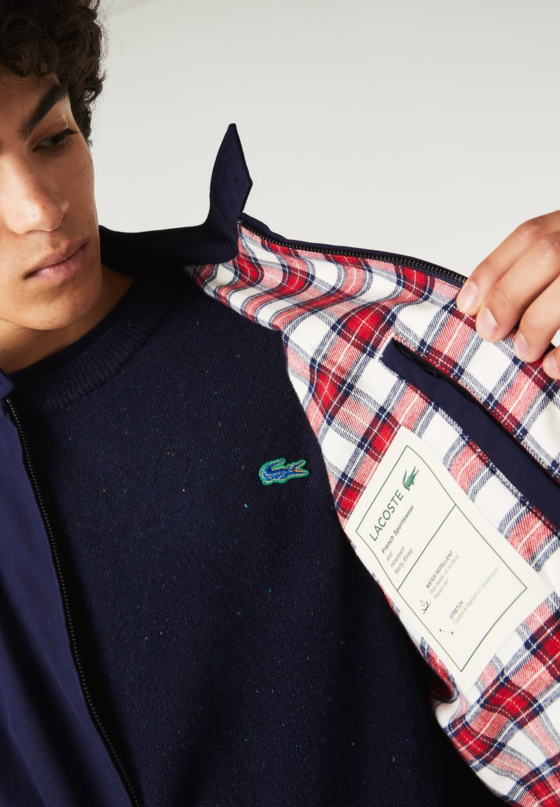 Veste navy avec un intérieur à carreaux comportant des motifs rouges, blancs et bleus ; comprend un logo brodé vert et des détails d'étiquette d'entretien à l'intérieur.