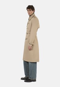 Trinchera beige con cintura ceñida y frente de doble botonadura. Presenta botones y hombreras. Combinada con jeans de pierna ancha y botas negras.