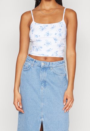Kvinde iført en hvid cropped tanktop med blåt blomsterprint og en lyseblå højtaljet denimnederdel med slidse foran.