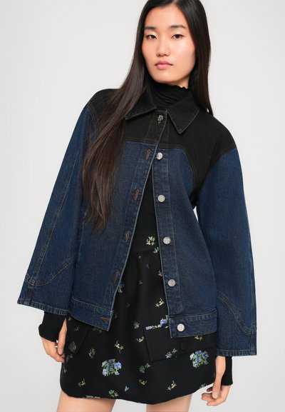 Traper jakna s crnim gornjim i tamnoplavim donjim dijelovima, sadrži srebrne dugmadi, široki ovratnik i opušteni kroj. Ispod je crna cvjetna haljina.