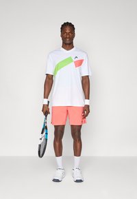 Hvid sports T-shirt med grønne og pink diagonale accenter, parret med koralfarvede shorts, hvide sokker og tennissko, mens der holdes en tennisketsj.