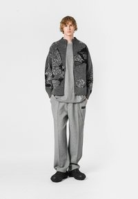 Cardigan de punto gris con patrones negros, superpuesto sobre una camiseta gris, combinado con pantalones deportivos grises y calzado negro texturizado.