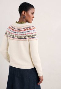 Suéter color crema con un canesú estampado que presenta motivos florales en rojo, verde y azul. Puños y dobladillo acanalados. Llevado con una falda oscura.