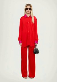 Κόκκινο πουκάμισο με κουμπιά και ροζ γιακά, συνδυασμένο με φαρδιά κόκκινα παν trousers. Το μοντέλο κρατά μια μικρή μαύρη τσάντα. Λείο ύφασμα, ελάχιστες λεπτομέρειες.