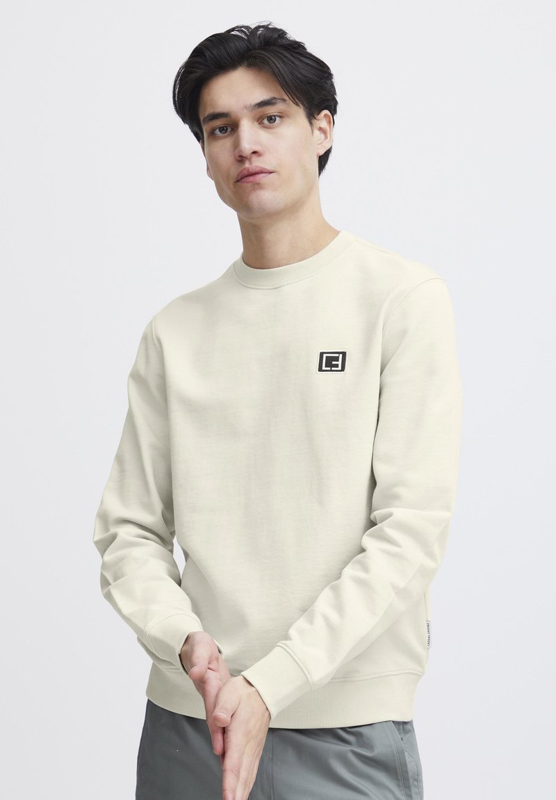 Hellbeige Sweatshirt aus weichem Stoff, mit Rundhalsausschnitt, langen Ärmeln und einem kleinen schwarzen Logodetail auf der linken Brust.