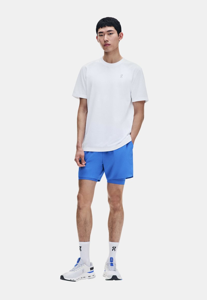 Jeune homme debout portant un t-shirt blanc, un short de sport bleu vif, des chaussettes blanches et des chaussures de sport blanches sur un fond uni.