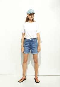 Blauwe denimshorts met omgeslagen zomen, gecombineerd met een eenvoudig wit T-shirt en een lichtblauwe pet. Zwarte sandalen met rode accenten.