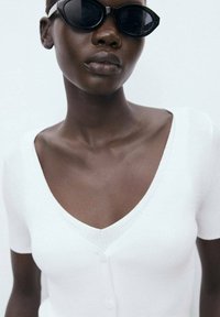 White ribbed short-sleeve top with a deep V-neckline and button detail, complemented by sleek black sunglasses against a neutral background.